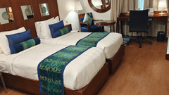 An angled view of the Superior Twin Room at Pride Premier Chennai displaying the two single beds in the foreground. A desk with an office chair and lamp is positioned along the back wall next to the window curtains.
