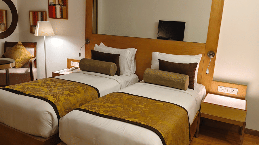 A front-facing view of the two single beds in the Deluxe Twin Room at Pride Premier Chennai. A bedside table sits between the beds, and a seating area with a mirror is positioned on the left.