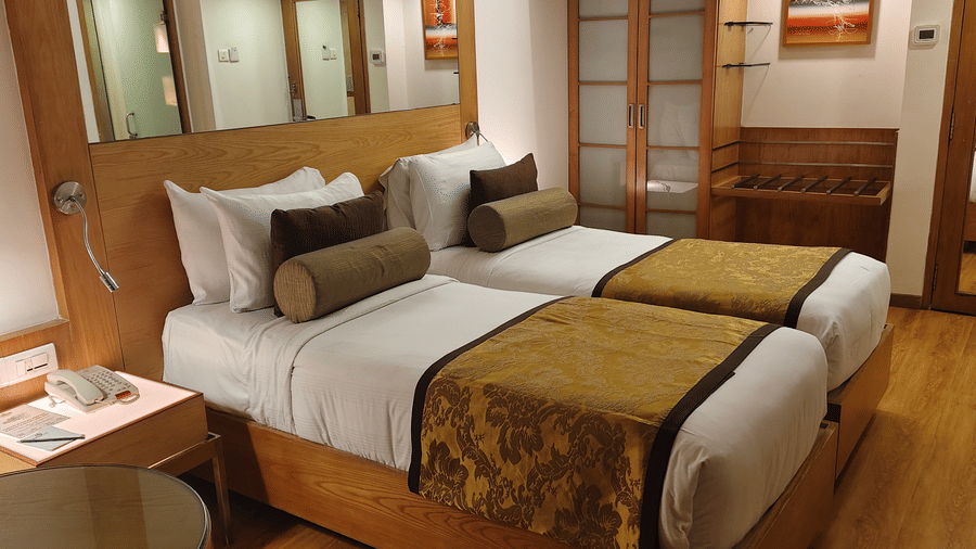 A view of two single beds in the Deluxe Twin Room at Pride Premier Chennai. A bedside table sits on the left, with the entrance and a mirror visible in the background.