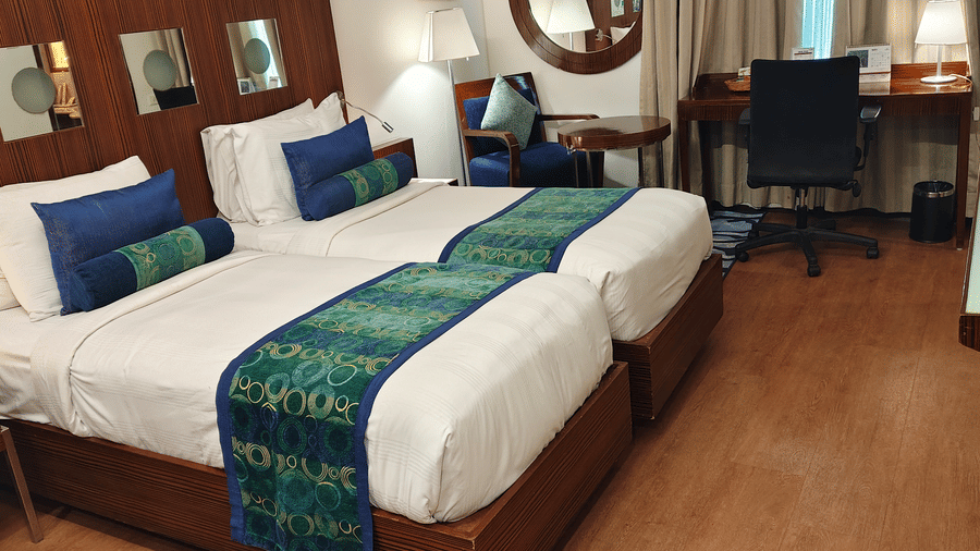 An angled view of the Superior Twin Room at Pride Premier Chennai displaying the two single beds in the foreground. A desk with an office chair and lamp is positioned along the back wall next to the window curtains.