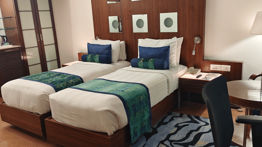 A view facing the beds and sitting area of the Superior Twin Room at Pride Premier Chennai. The wardrobe is visible on the left side, and a table with a chair sits on a rug near the right side of the room.