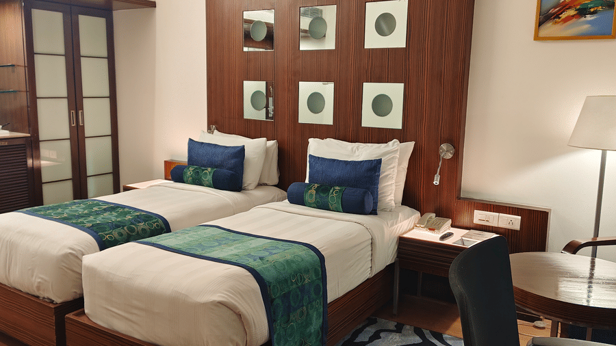 A wide view of the Superior Twin Room at Pride Premier Chennai showing two single beds side by side. A wardrobe stands near the entrance on the left, and a lamp with a round table and chair sits to the right of the beds.