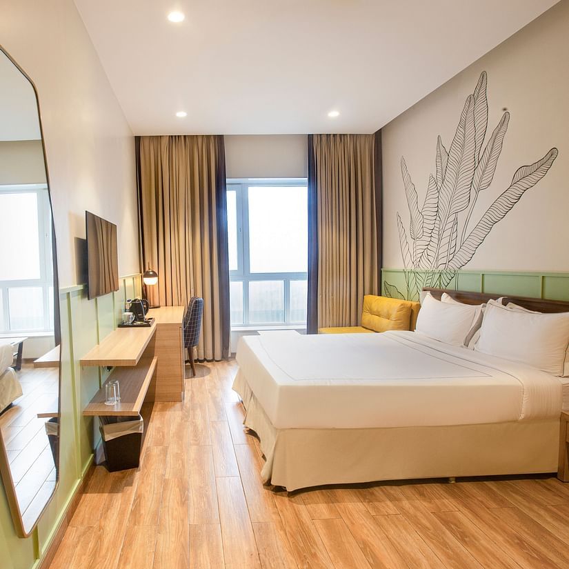 A side wide-angle view of the Superior Deluxe room at Hotel Bawa Regency, Dadar, featuring a comfy bed, a large hexagon-shaped mirror, a flat-screen TV, and a balcony.