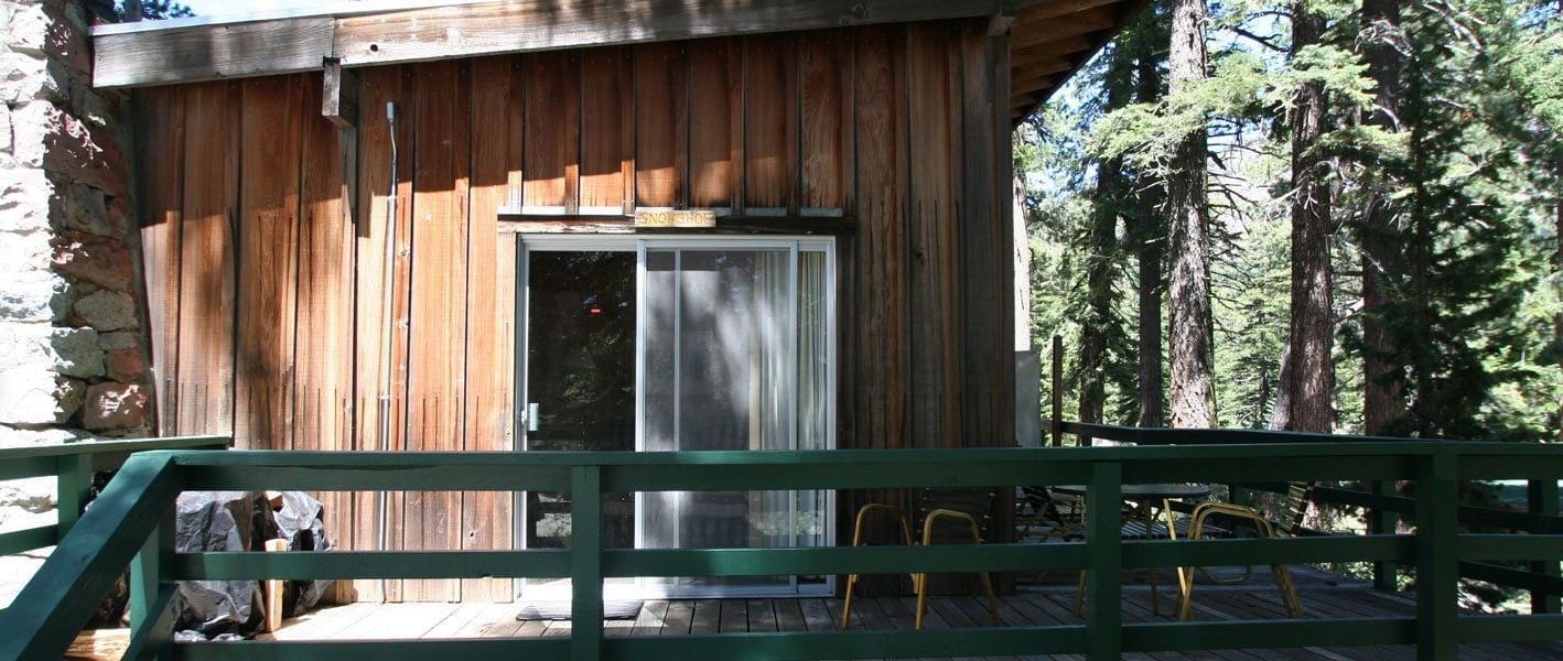 A rustic wooden cabin with a slanted roofline, featuring a stone chimney and a sliding glass door. - Kit Carson Lodge