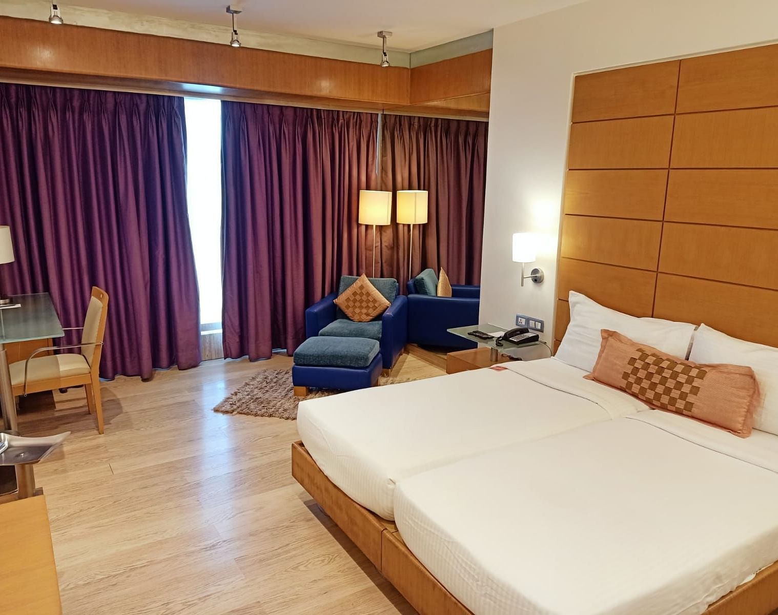 Image of twin beds in the executive twin room at our hotel featuring a flat screen television, bed side table, mirror, violet curtains, and other furniture. | Mirage Hotel