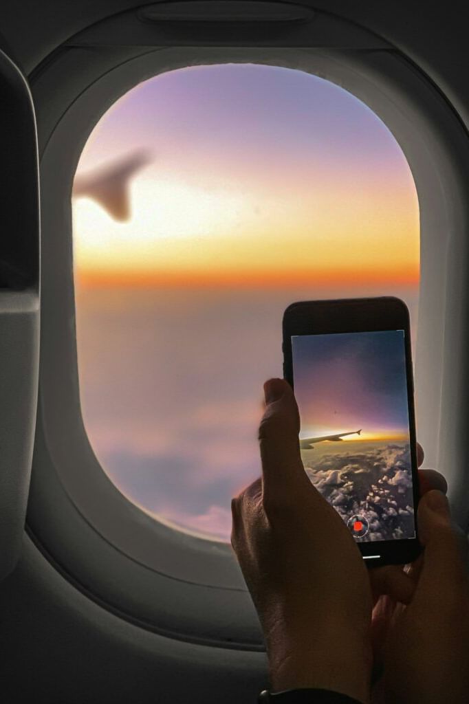 A hand holds a smartphone capturing a sunset view from an airplane window.