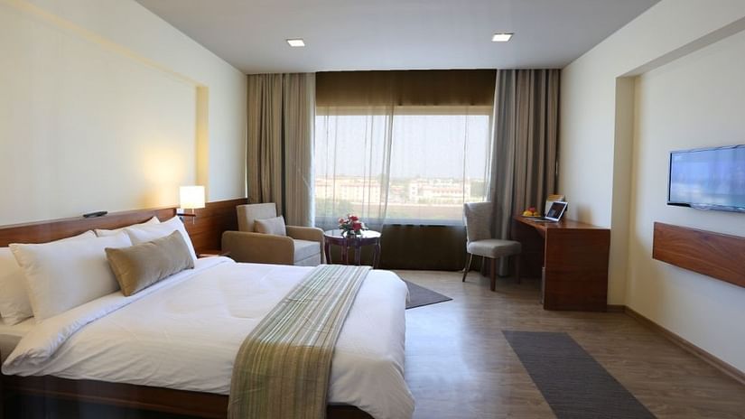 A bed room with a large, well arranged bed with mattress, facing a flat screen television, with a view to the cityscapes 