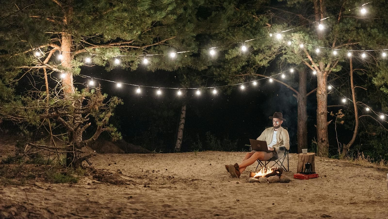 A wide-angle image of a forest area illuminated by warm lights, featuring a man sitting with a laptop next to a bonfire, with a car parked nearby.