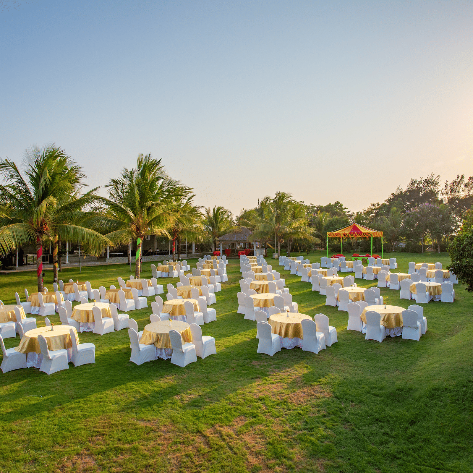 An outdoor event space on a green lawn with numerous round tables and chairs, set up for a gathering under a bright sky surrounded by palm trees - Grande Bay Resort & Spa, Mamallapuram
