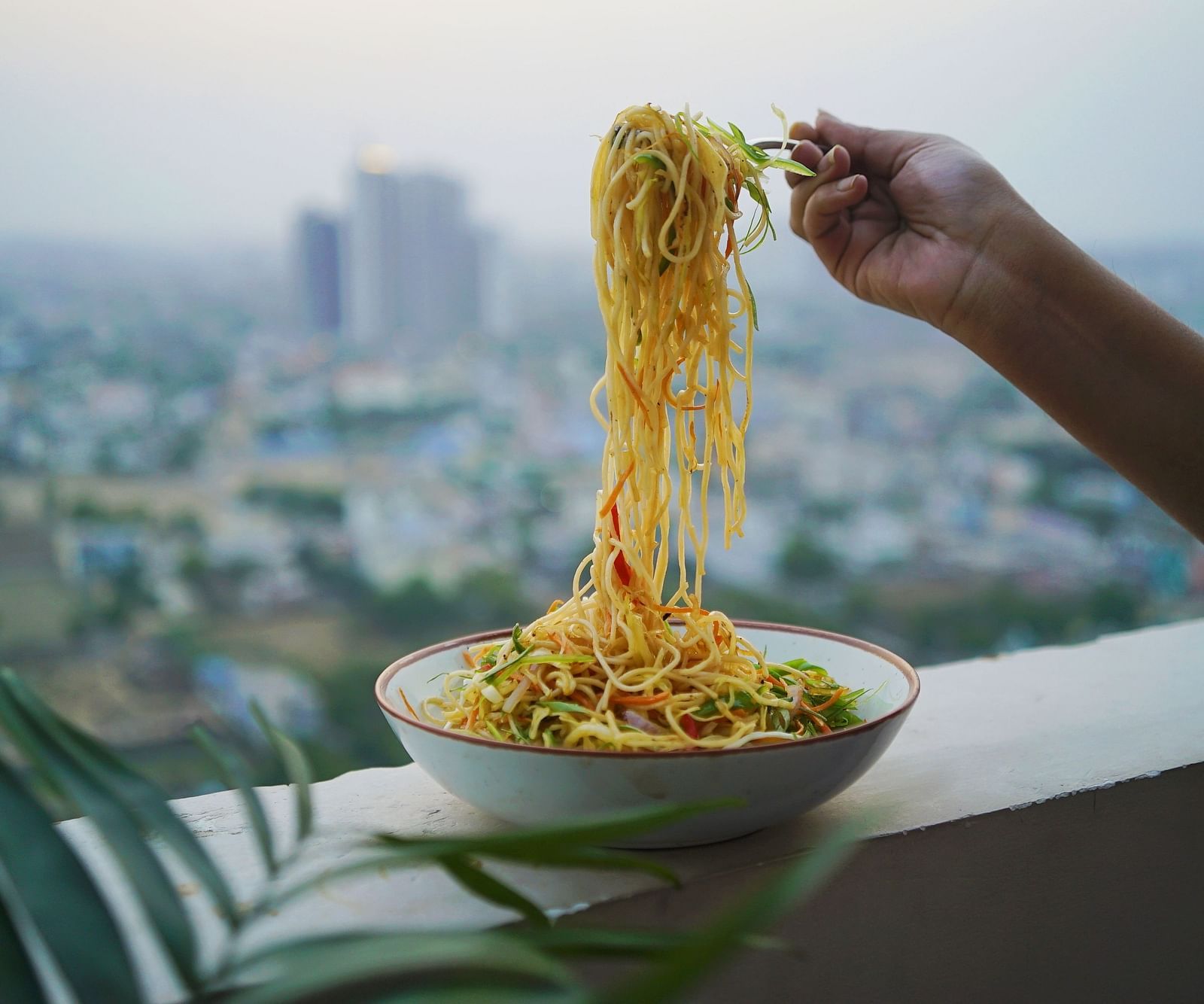 A bowl of noodled kept on the ledge with a person taking a handful from a fork - Rhythm Gurugram