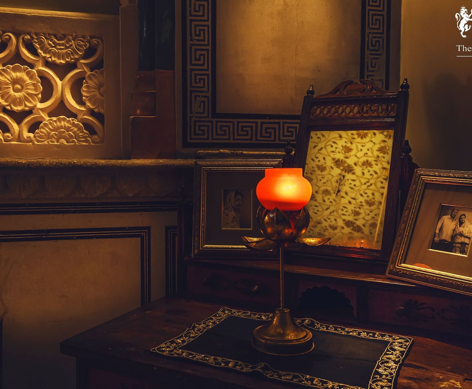 A dimly lit room at Umaid Vilas Jaipur featuring a decorative wall cutout, a lamp with an orange shade, and framed artwork.
