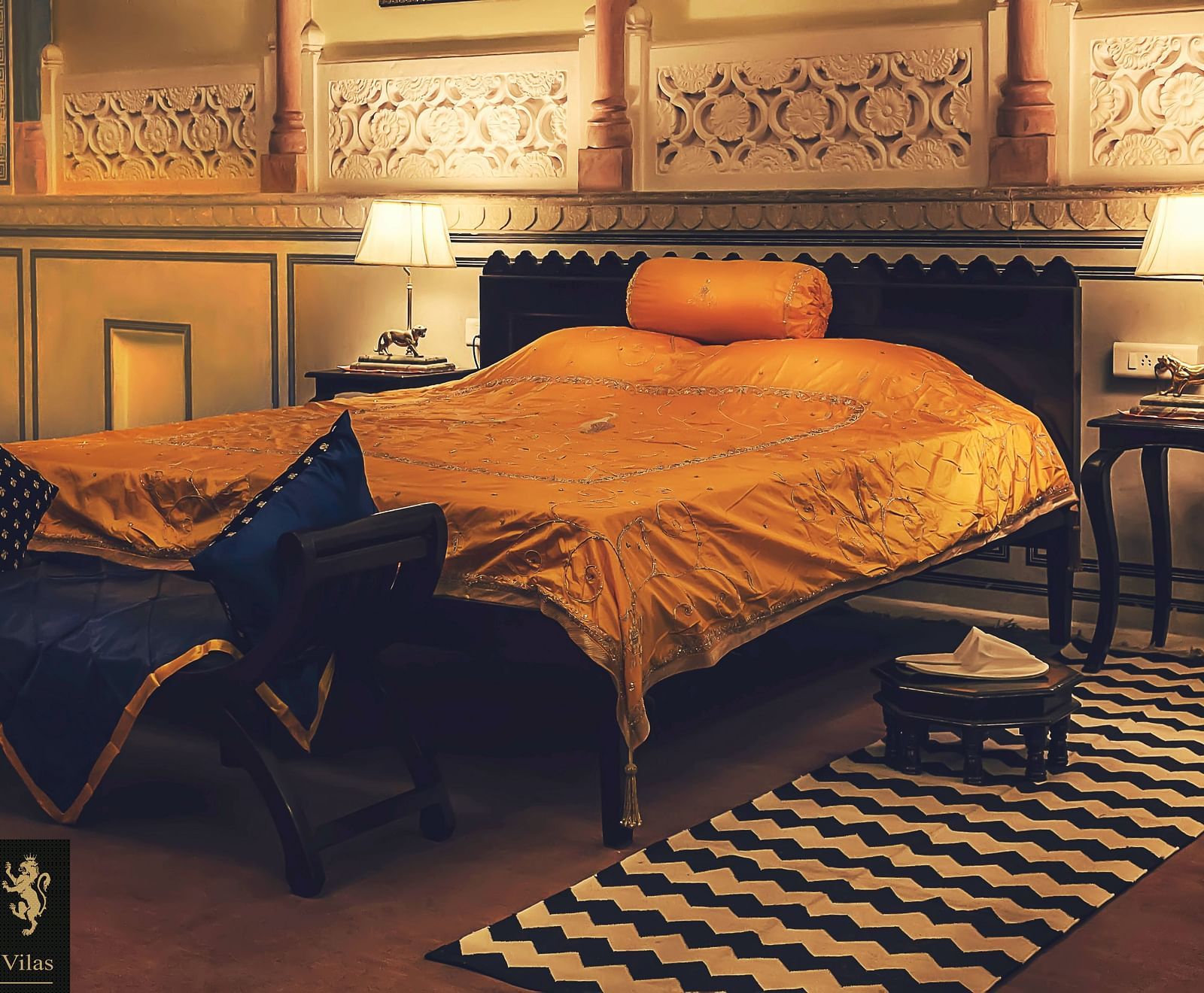 A bedroom at Umaid Vilas Jaipur with a bed dressed in golden fabric, two lamps, and decorative wall cutouts.