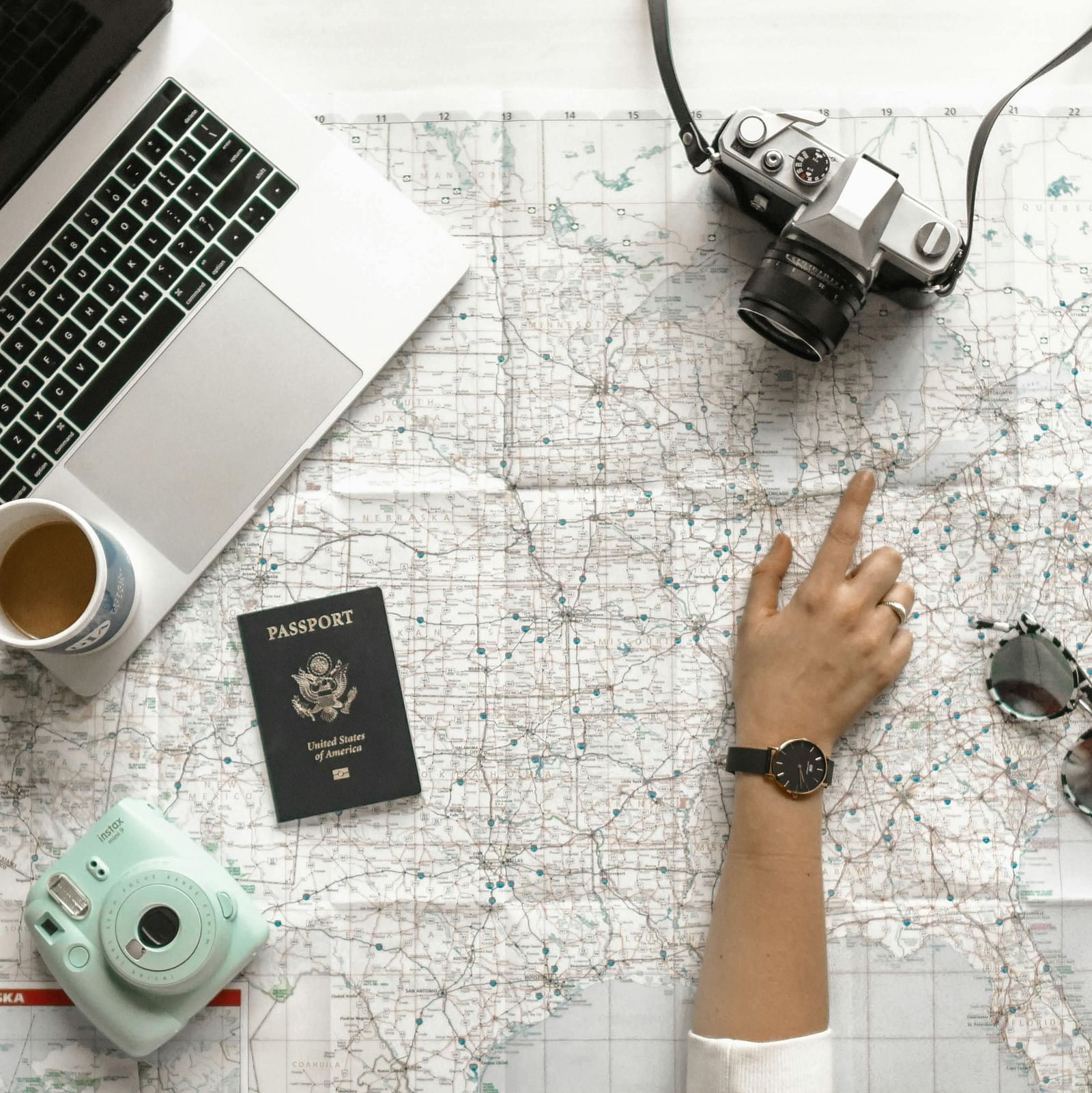 A person pointing a finger at the map on the table surrounded by sunglasses, a digital camera, a laptop with coffee mug on it, a polaroid camera and a passport