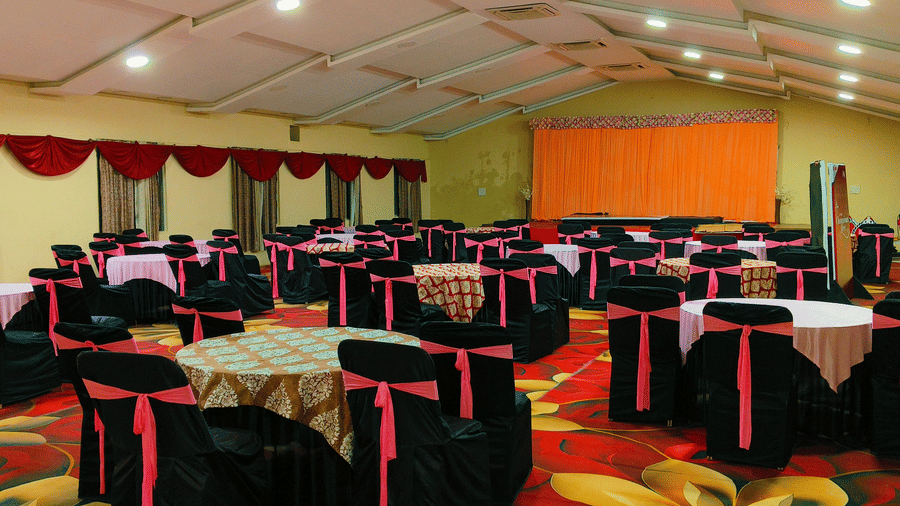 An indoor banquet hall with a high white tent-like ceiling, set up with round tables surrounded by chairs covered in black and red cloths, and a curtained wall in the background | Nandan Resort