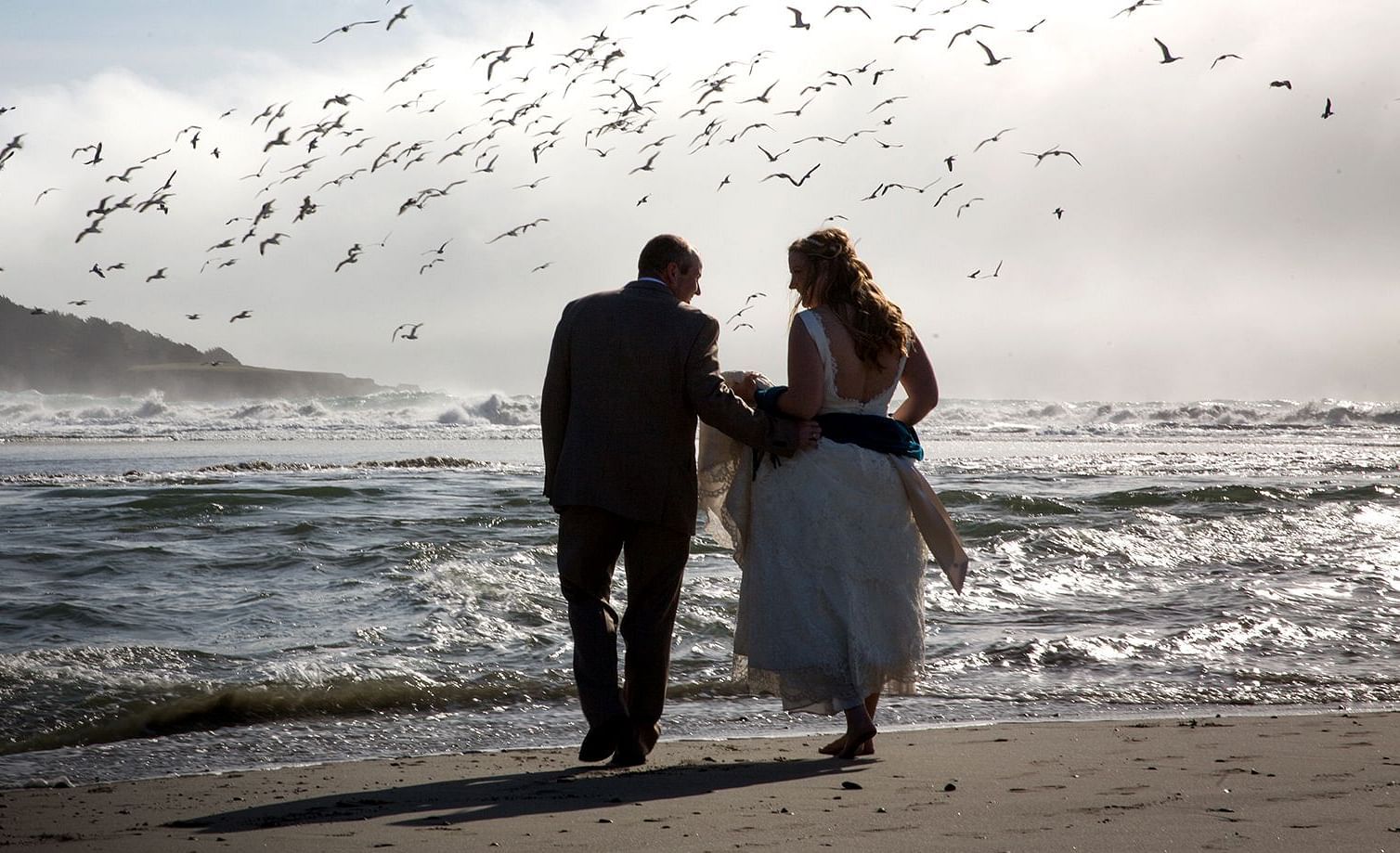 Newlyweds strolling on the beach as seagulls take flight around them