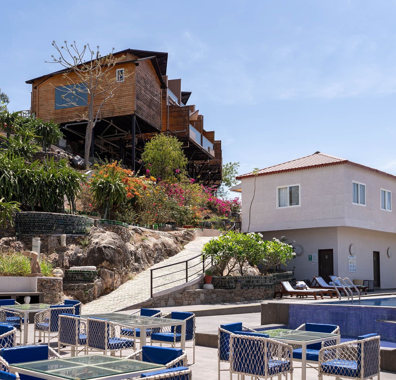 A multi-level building on a hill or slope and has a rustic or traditional look. In the foreground, there's a patio or terrace with outdoor furniture, including tables and chairs - Bamboo Saa Resort & Spa, Udaipur