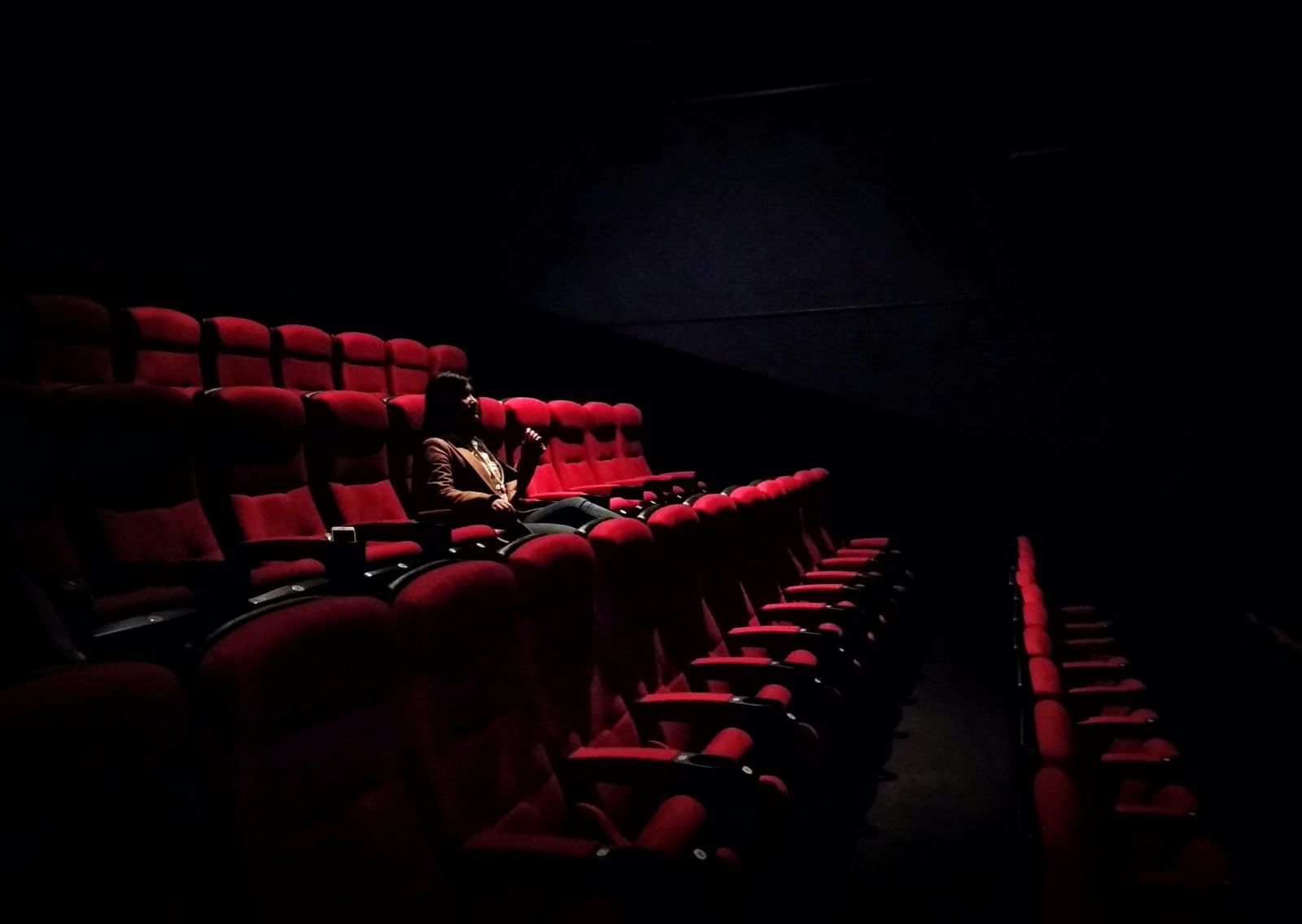 A solitary woman sitting in a movie theatre consisting of red chairs and a spotlight on her.
