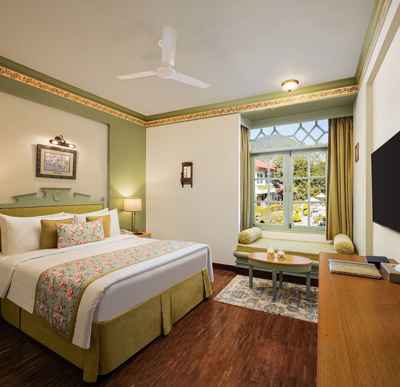 An image of a  cozy room in the naini retreat, nainital, with a king-size bed, wooden furniture, and a window. The room also features a ceiling fan, a TV and wooden flooring.