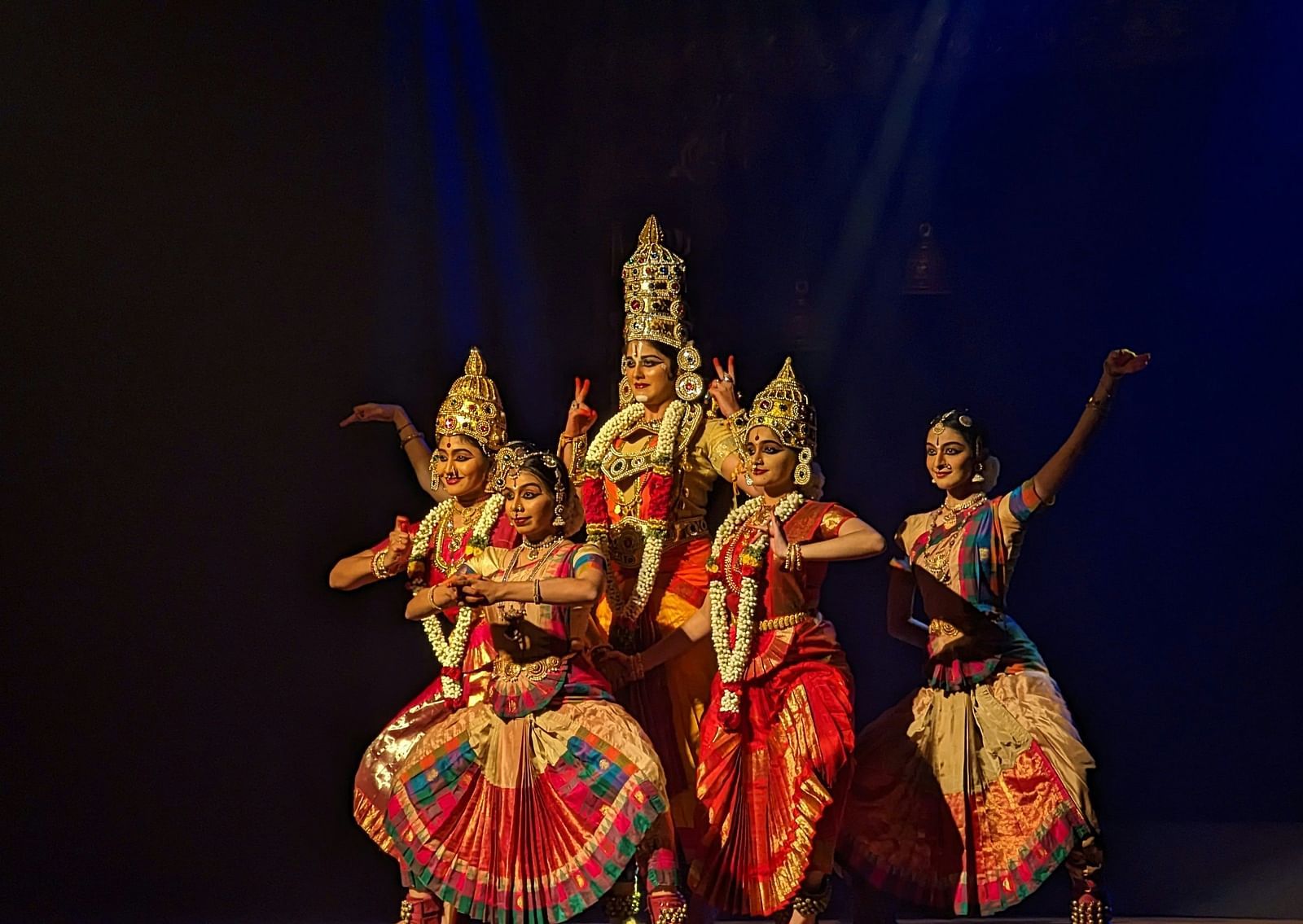 A group of women performing bharatanatyam on stage with a black background.
