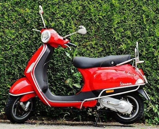 A vibrant red Vespa scooter is parked against a lush green hedge.