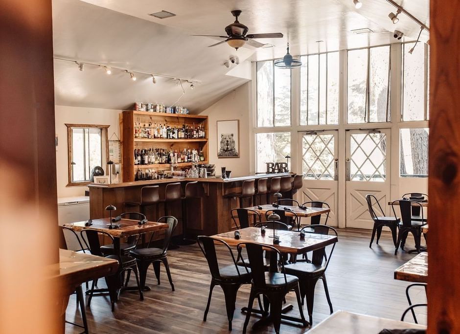 A cafe at Shaver Lake Village Hotel with wooden tables, metal chairs, large windows, and warm lighting, creating an inviting ambiance.