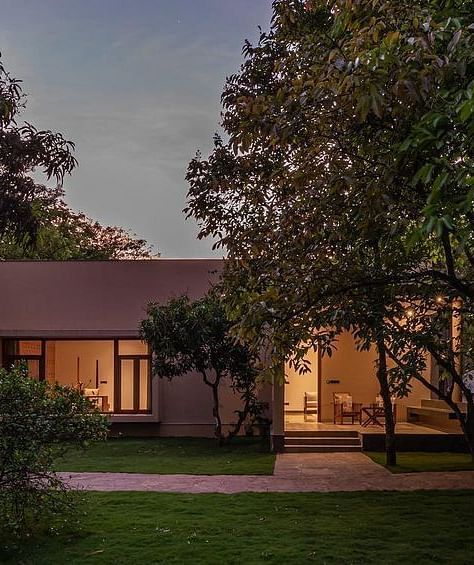 An exterior view of a building with lit windows, surrounded by trees and greenery at dusk - Shreyas Retreat