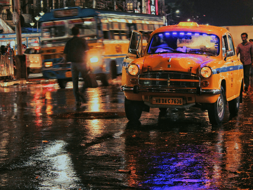 A vintage yellow taxi driving through a rainy city street at night with reflections on the wet road.