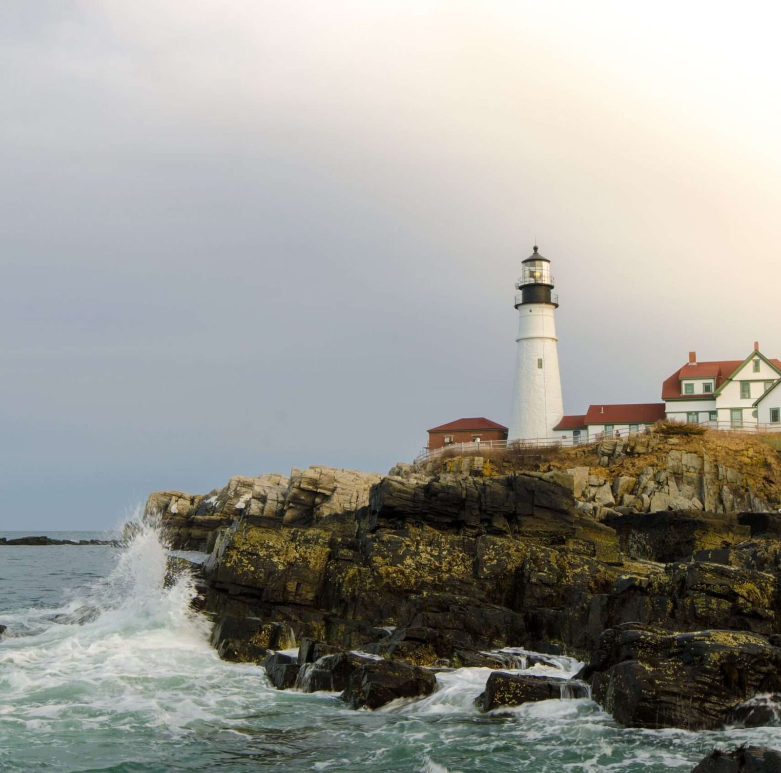 A wide angle shot of a light house at the end of a cliff with waves crashing against the shore under a stormy sky.
