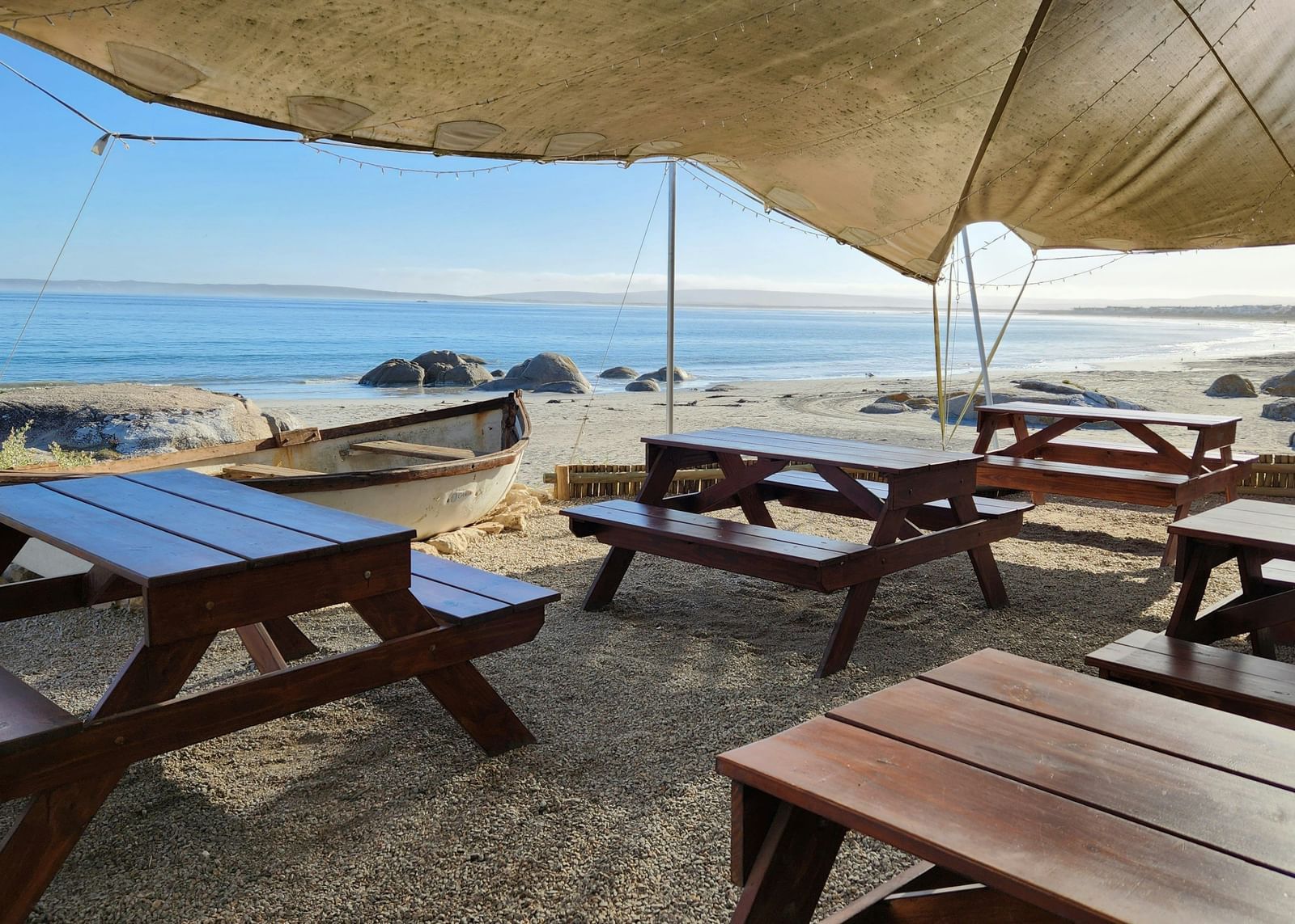 A view of a beach-side restaurant with a canopy above and wooden tables below it.