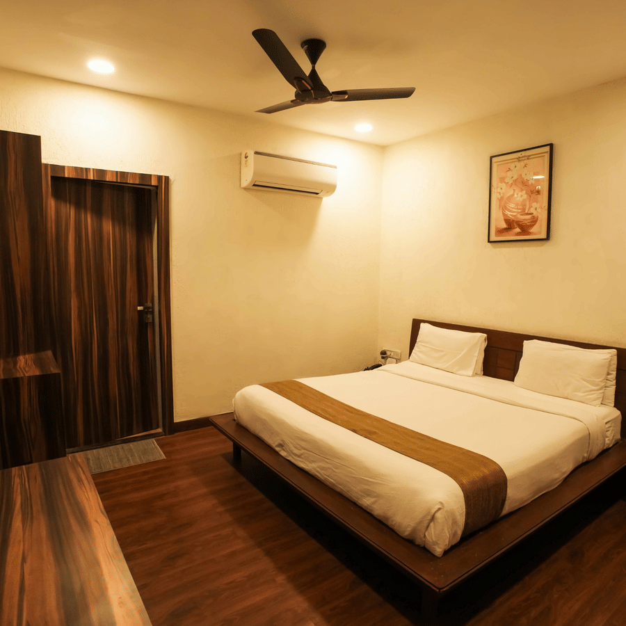 A view of bedroom at Adamo The Village with plush bedding, wooden flooring, a flat-screen television, and warm lighting.