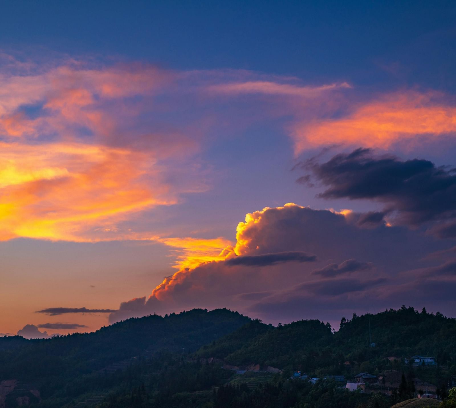 A vibrant sunset sky with shades of orange, purple, and blue, clouds lit by the sun and silhouetted mountains in the foreground.