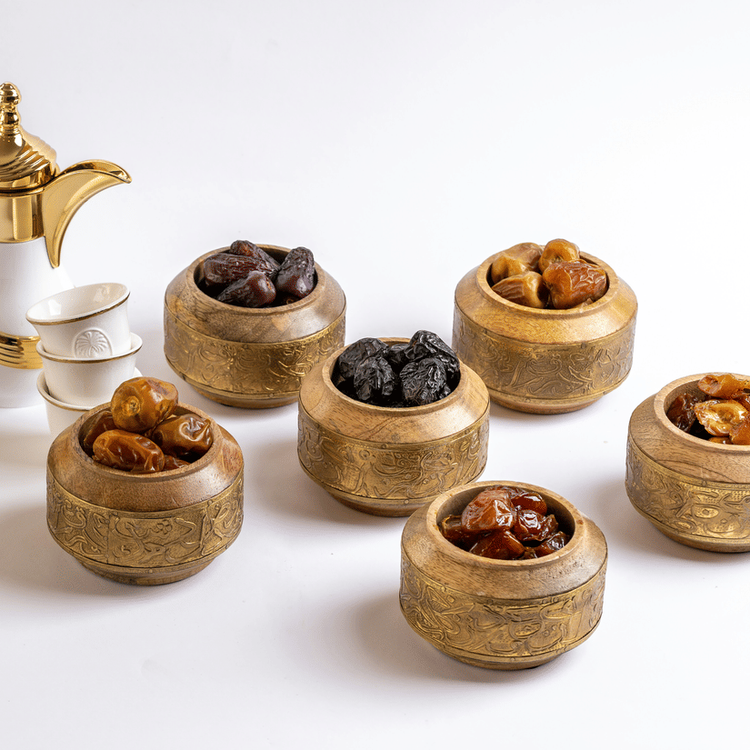A view of numerous types of dates kept in a brass cup next to each other with a pot of Dallah Coffee next to it.
