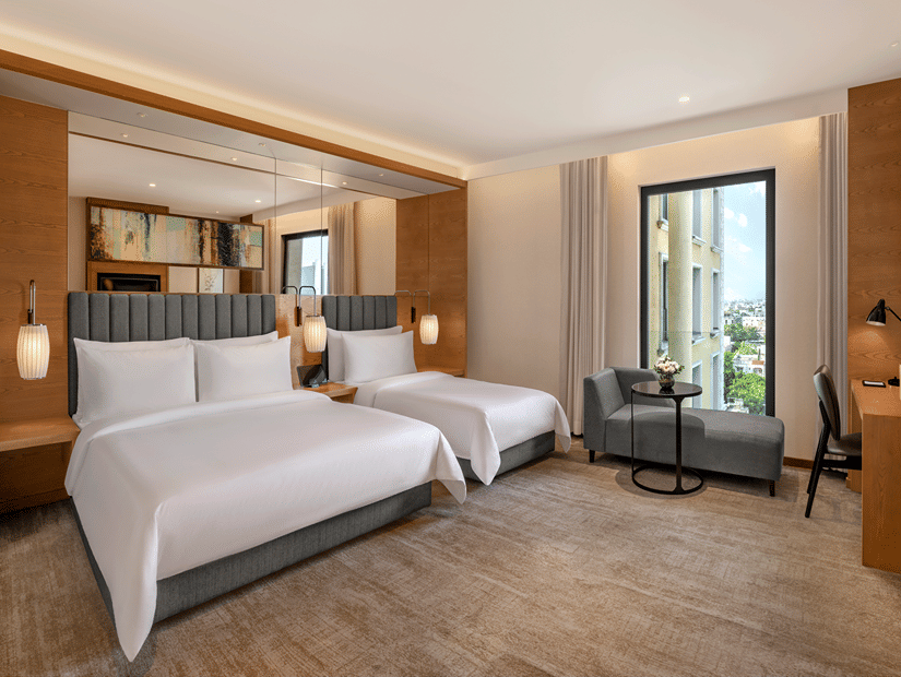 Twin beds with white bedding and grey headboards in a room at The Residency Towers Chennai, with wood panelled walls, a grey armchair and a window view.