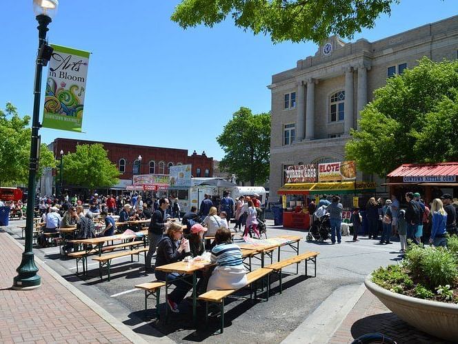 A lively street with people dining at picnic tables, enjoying food, drinks, and a vibrant social atmosphere.- The Grand Hotel & Ballroom, McKinney