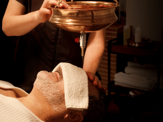 A close-up shows a man lying down with a towel on his forehead, receiving a shirodhara treatment with warm oil pouring from a brass pot at YO1 Longevity & Health Resorts