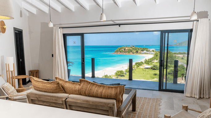 A luxurious living room with a large window offers a panoramic view of a turquoise ocean and a distant beach.