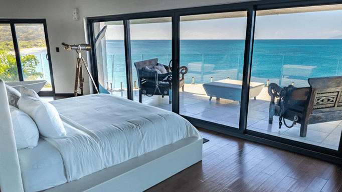 A bedroom with a large bed faces expansive glass doors that open to a panoramic view of the ocean.