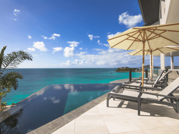 A resort swimming pool with lounge chairs is situated next to a lush landscape and overlooks the ocean.