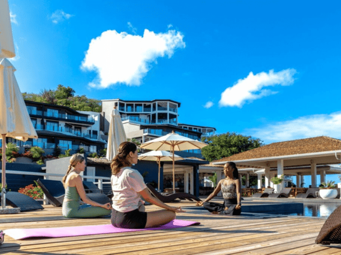 A person practices yoga on a wooden deck overlooking a sunny beach with resort buildings in the background.