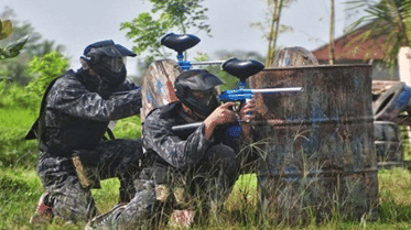 Two guests in black protective gear and masks crouching behind a wooden barrier during an outdoor paintball activity at Vedic Village Spa Resort.