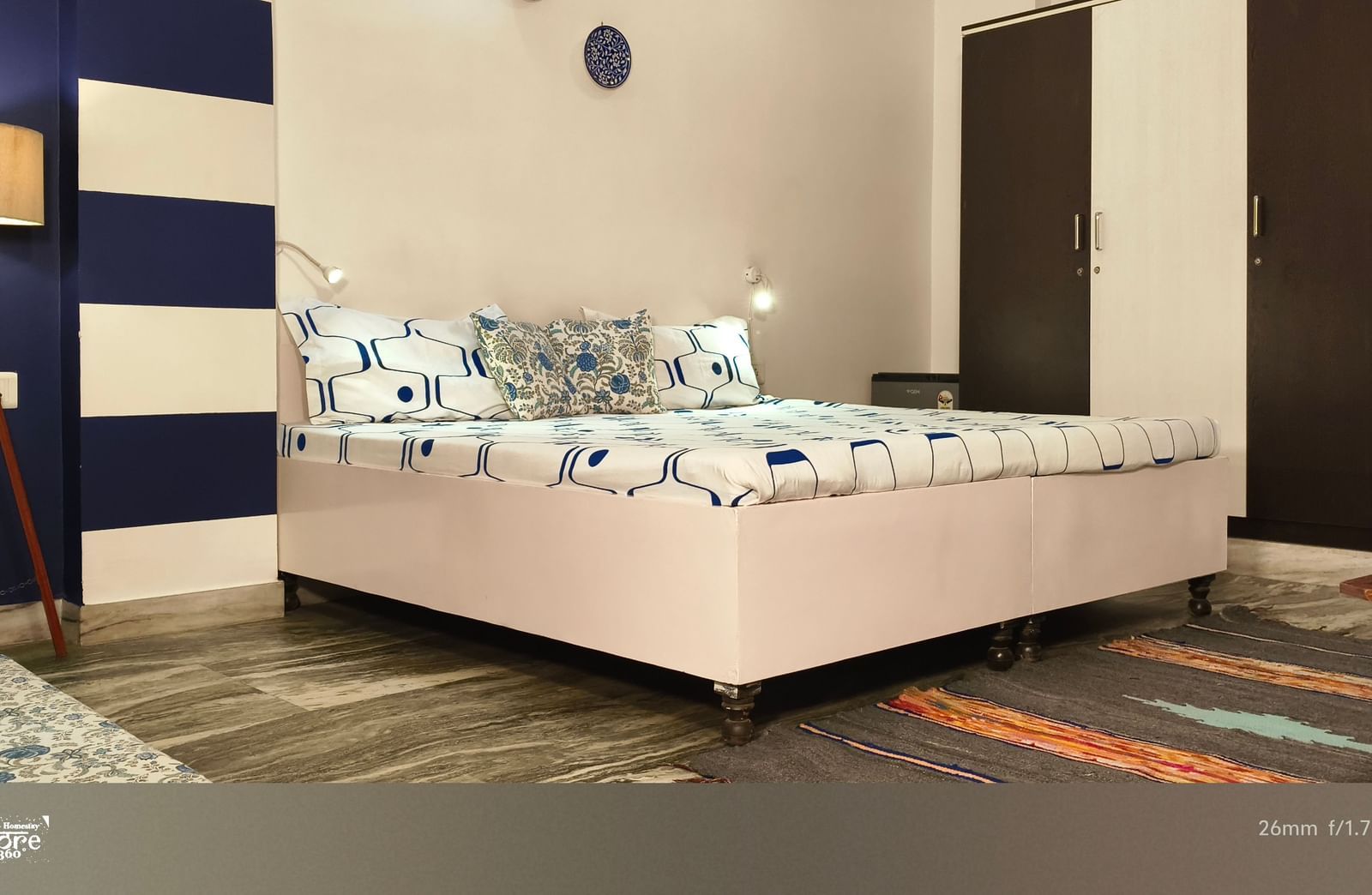 A neatly arranged bedroom with a double bed, blue accent wall, bedside lamp and rug on the floor at Jaypore360 Homestay - Best Rooms in Jaipur.