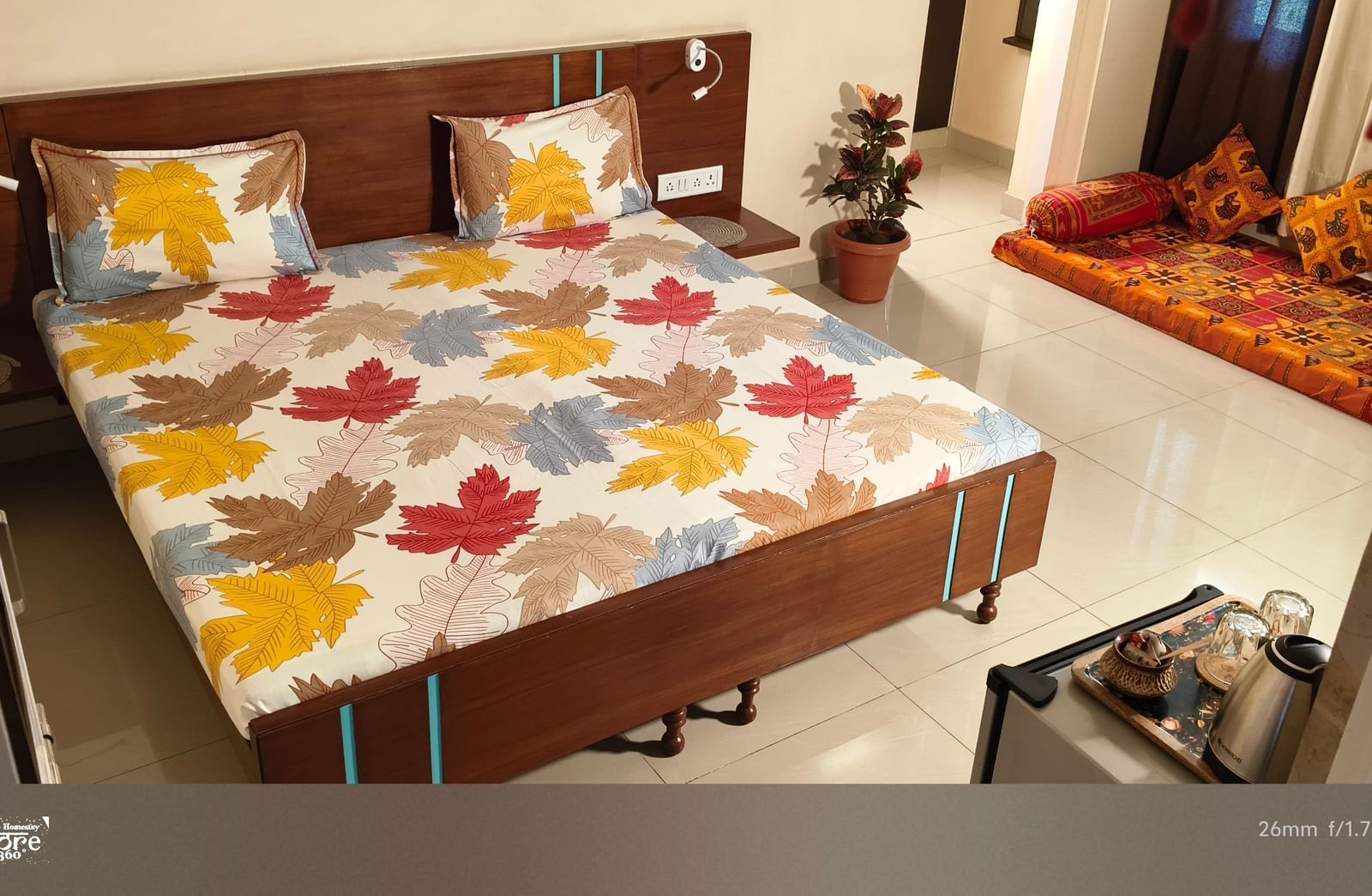 A bedroom with a double bed, floral bedsheet, side tables and a small seating area with cushions at Jaypore360 Homestay - Best Homestays in Jaipur.