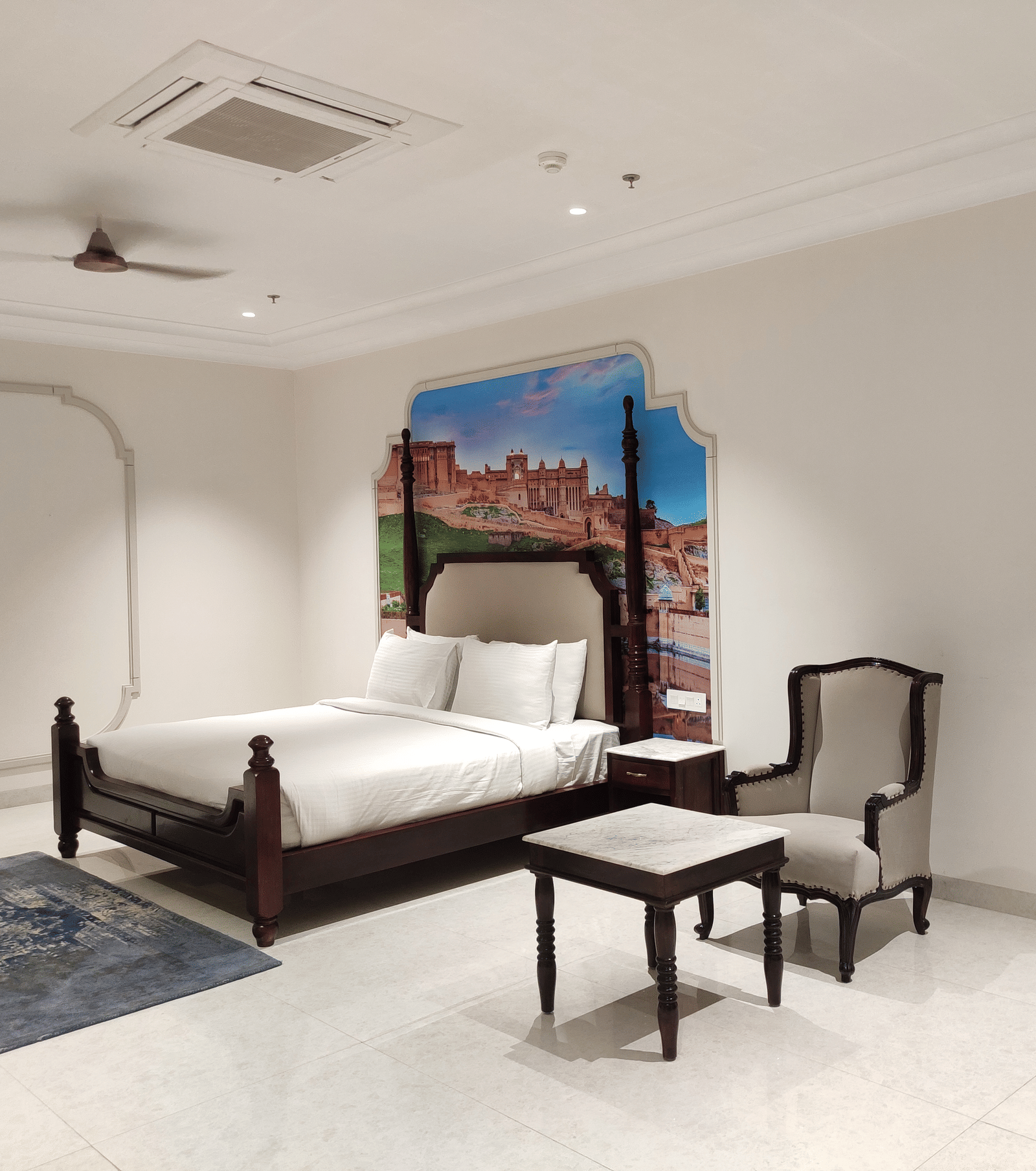 A bedroom at Anant Mahal, Jaipur featuring a king size bed, a small wooden table with a chair, a decorative mural on the wall, a ceiling fan and an A/C.