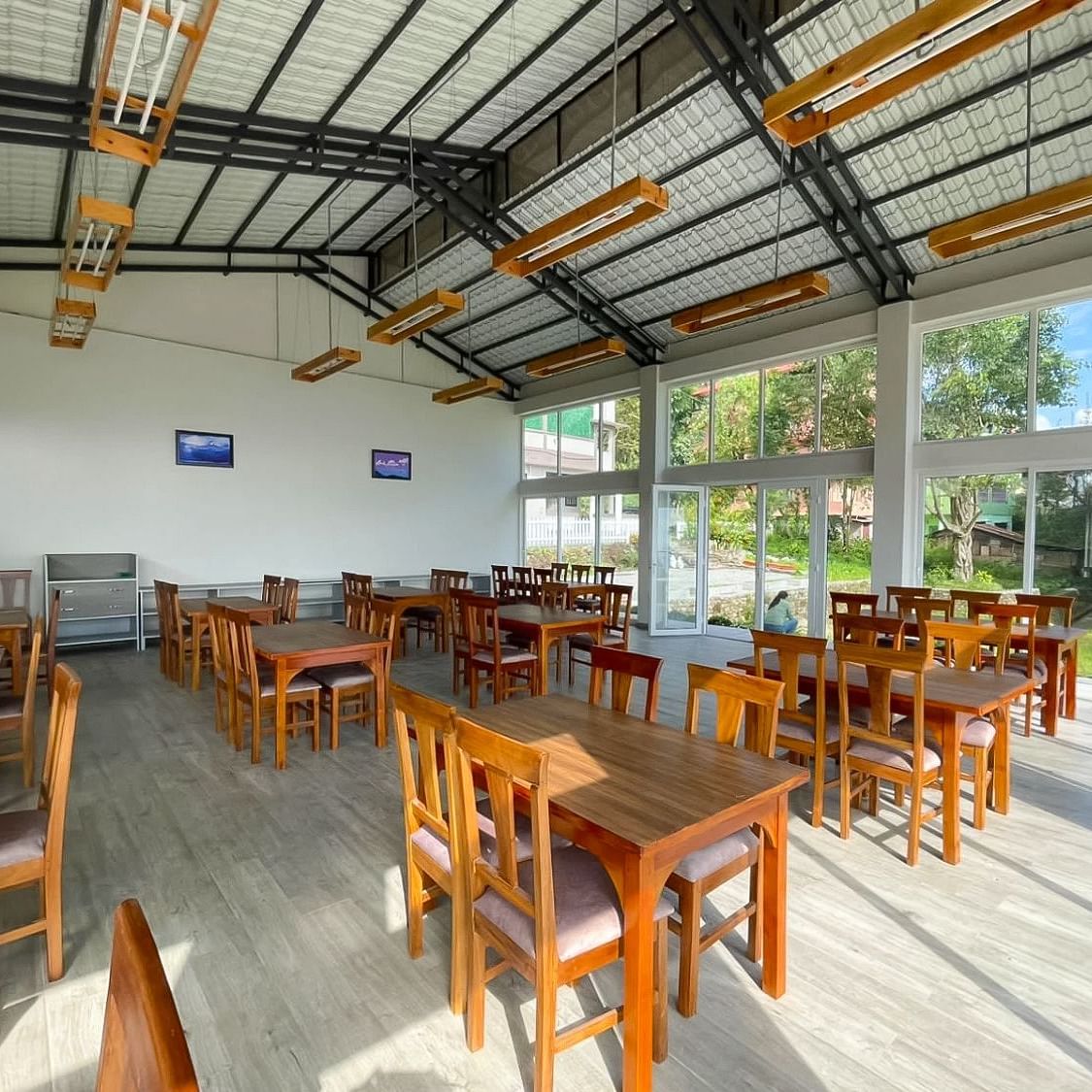 A spacious and bright dining area at Yashshree Resorts & Spa, Kalimpong, featuring numerous wooden tables and chairs, under a high ceiling with exposed beams.