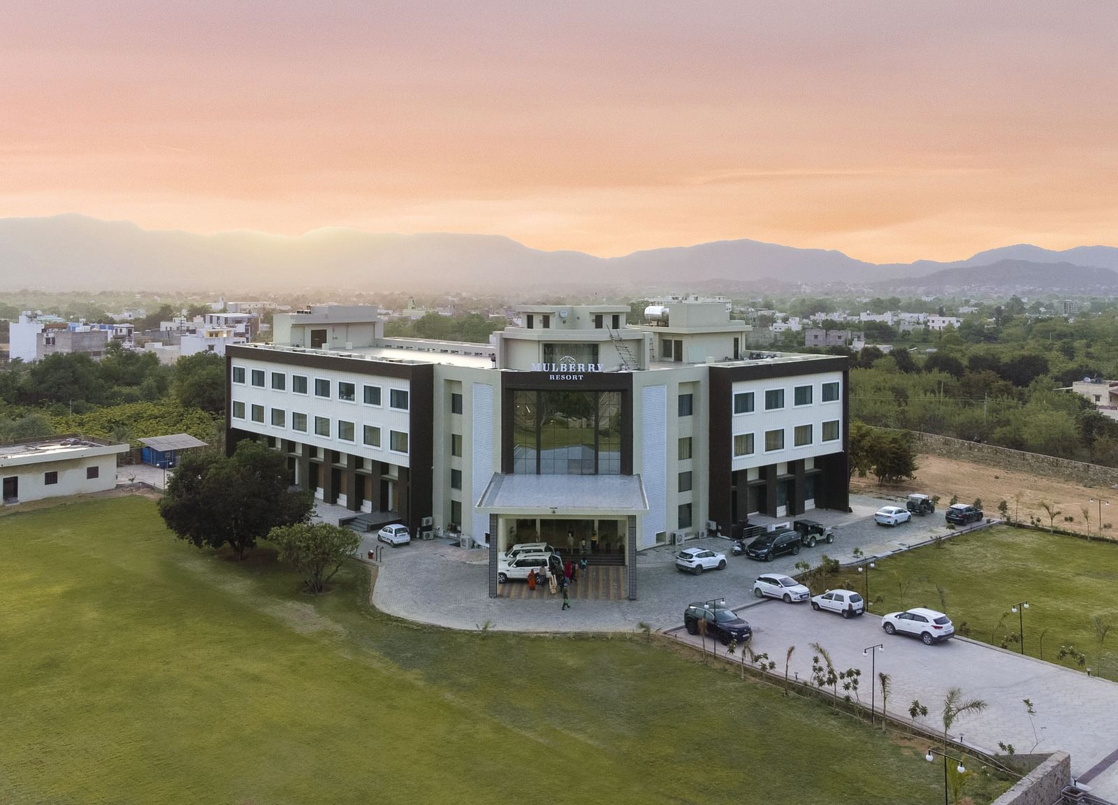 An aerial view of the Bamboo Saa Mulberry Resort, Pushkar at sunset or sunrise surrounded by green lawn and the backdrop of hills.