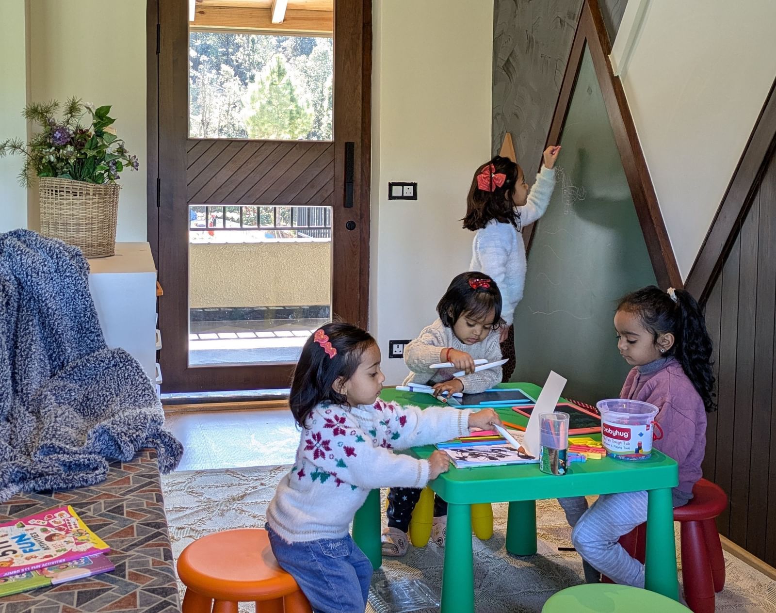 Spacious room with three kids seated around a small table playing, and a child writing on the board on the wall at The Chamomile Estate.