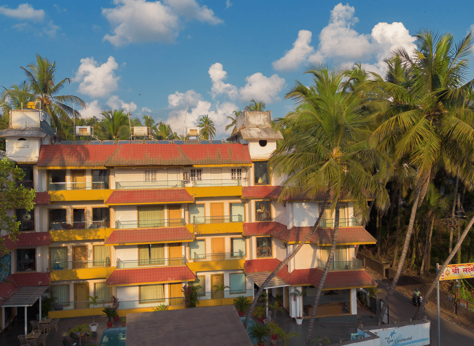 A colourful, low-rise hotel complex with many windows, surrounded by lush green palm trees under a bright sky - Perfectstayz Premium Goa Continental