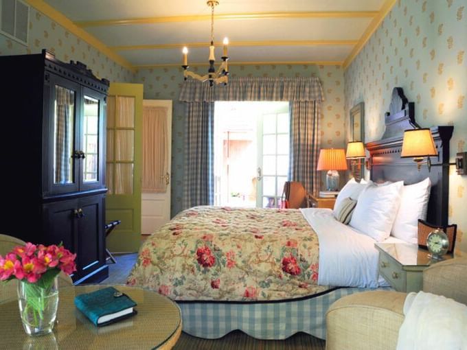 Side view of the Lower Garden Room at Tallman Hotel showcasing it's bed with floral linens, facing a vintage wardrobe in a room with a window and a seating area.