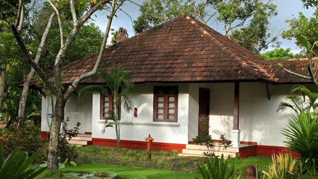 A single-storey, white colonial-style building with a distinctive deep red tiled roof, framed by lush tropical greenery and trees.