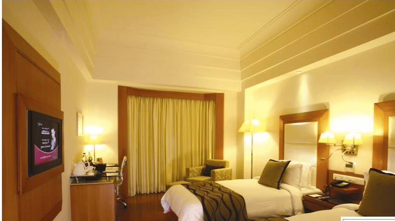 Deluxe Room Twin Bed at Ambrosia Sarovar Portico, Haridwar, featuring twin beds, bedside tables, a TV, work desk with chair, curtains, and wooden flooring.